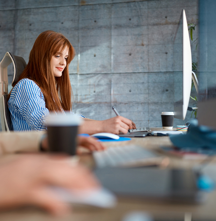Smiling woman with red hair working at a desktop computer, taking notes in a bright office with coffee cups nearby