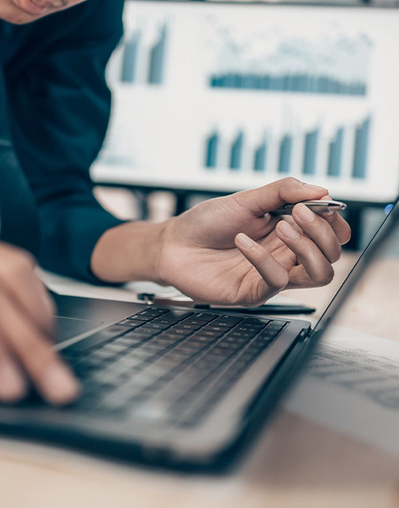 Close-up of a person using a laptop with financial graphs displayed on a screen in the background