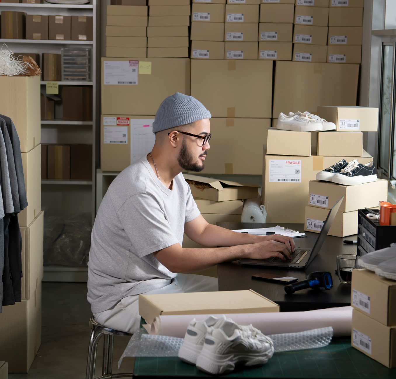 Man in a hat and glasses working on a laptop in a clothing webshop, surrounded by boxes and shoes